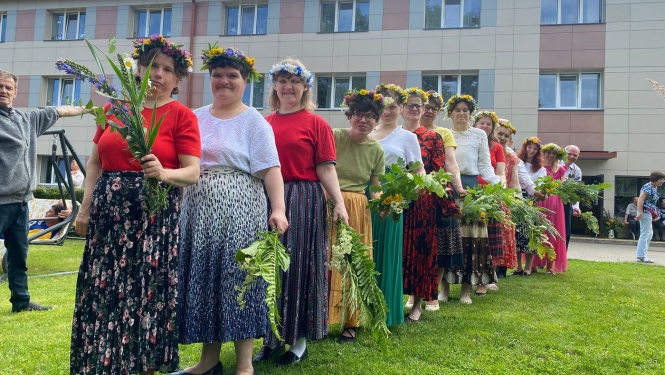 Svinīgi ģērbušās dāmas ar vainagiem galvās un jāņu zālēm rokās. Stāv rindā viena aiz otras un gatavojas uzstāties.