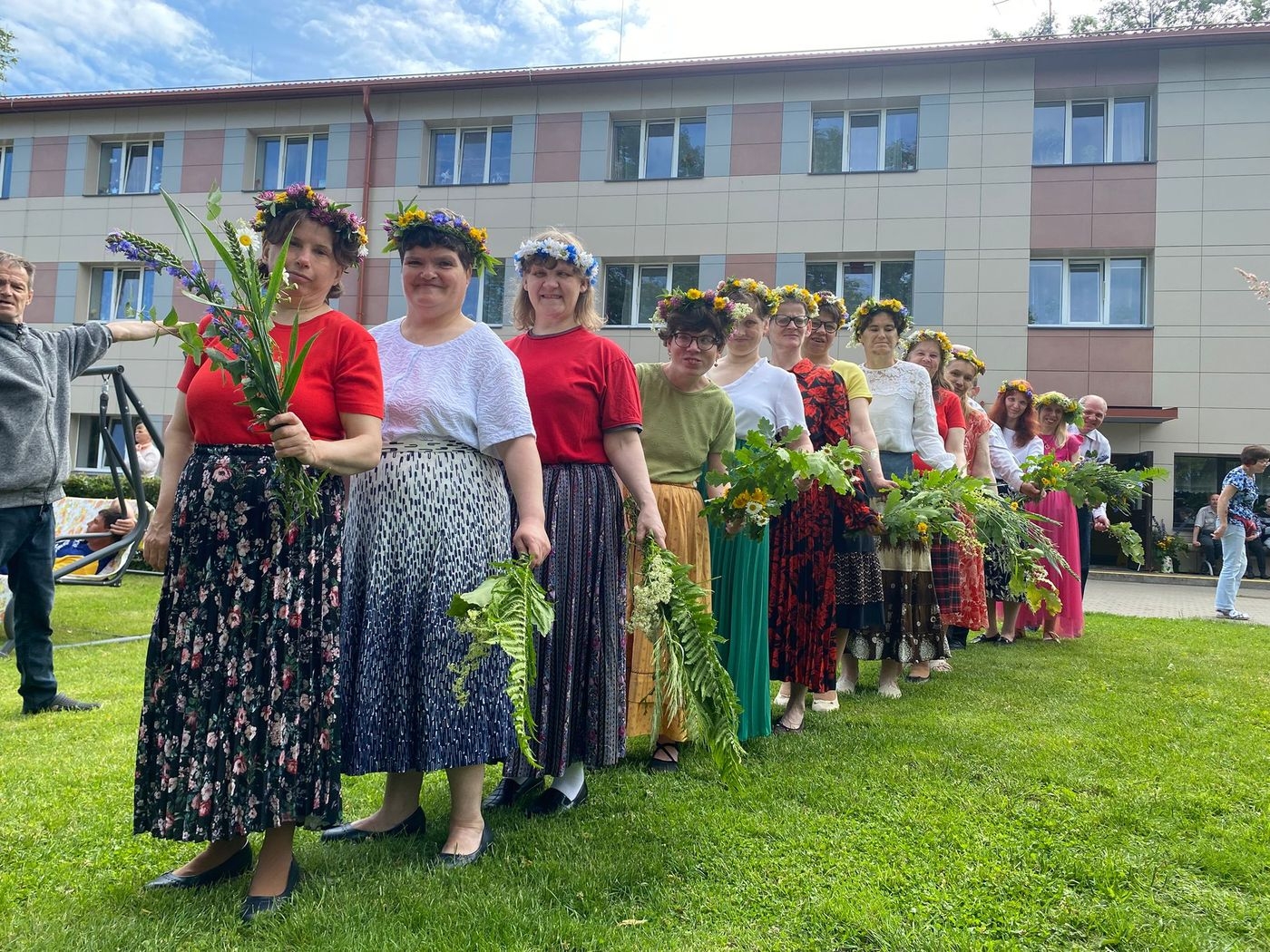 Svinīgi ģērbušās dāmas ar vainagiem galvās un jāņu zālēm rokās. Stāv rindā viena aiz otras un gatavojas uzstāties.