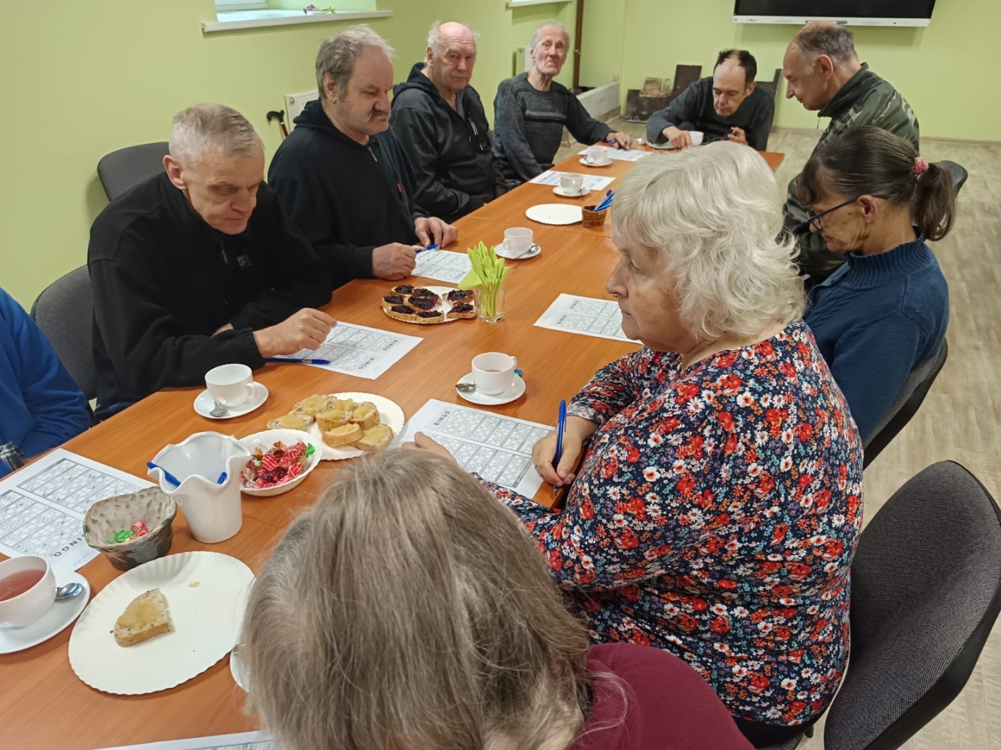 Pasākuma dalībnieki pie galda dzer tēju ar cepumiem un spēlē bingo
