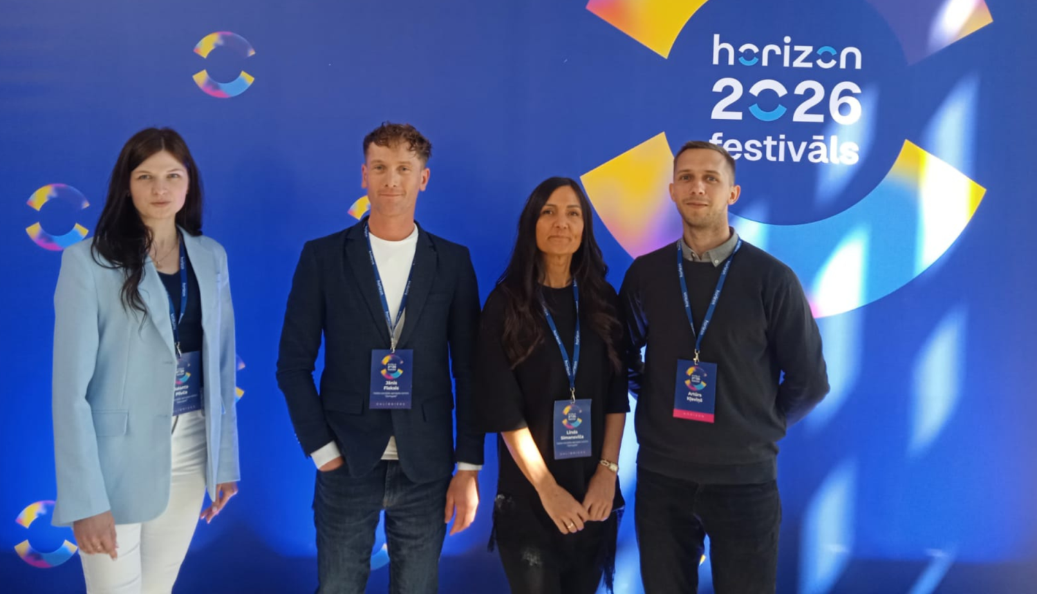 Četri festivāla dalībnieki uz koša fona banera