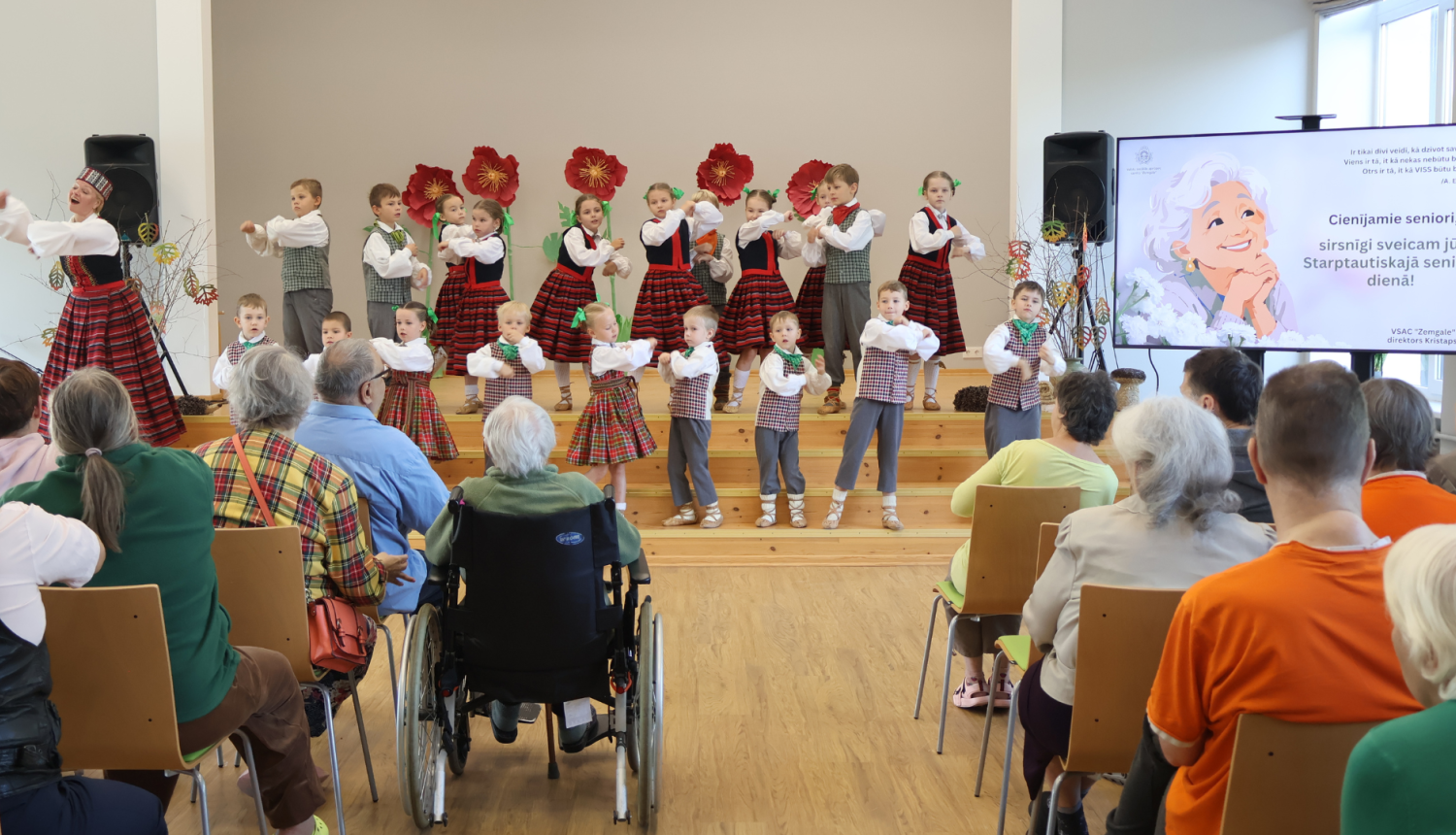 Bērni tautastērpos dejo uz skatuves. Viņus vēro spkatītāji