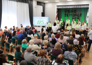 Uz skatuves iedzīvotāju apvienotais koris, baltajos kreklos, melnajās biksēs un visiem zaļas zīda šalles. 