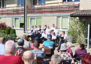 Skatītāji vēro svētku koncertu. Priekšā ansamblis baltos kreklos dzied