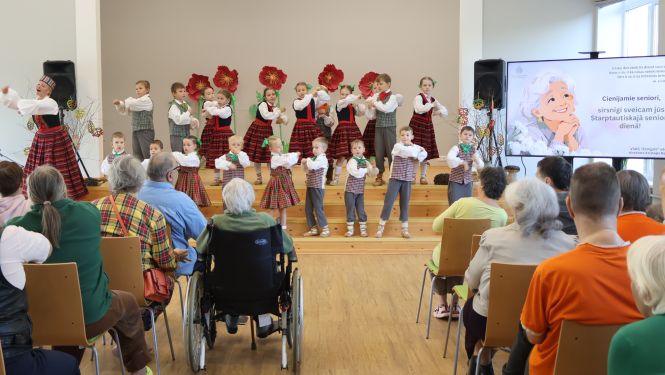 Bērni tautastērpos dejo uz skatuves. Viņus vēro spkatītāji