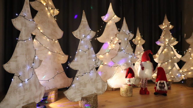 Attēlā skatuves dekorācijas. Trīs rūķi sarkanajos tērpos un rinda ar baltām eglēm, rotātā ar gaismiņām.