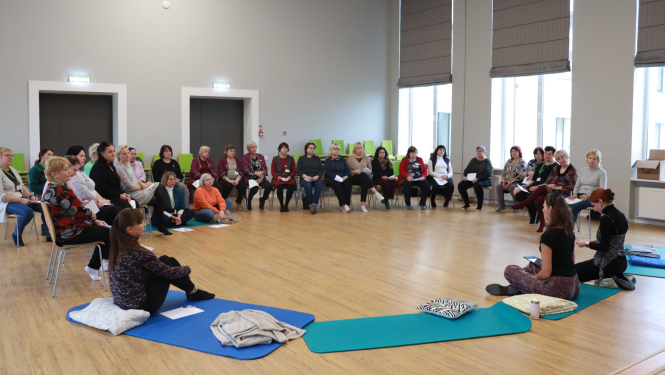 Pasākuma dalībnieki zālē sēž krēslos, kuri izkārtoti aplī, priekšā divas mākslas terapeites vada nodarbību