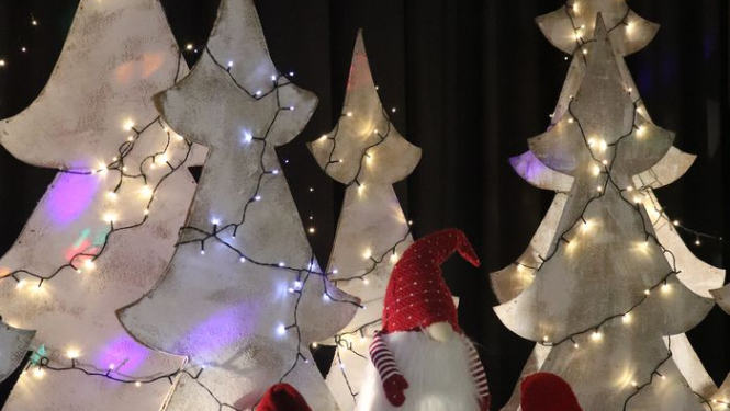 Attēlā skatuves dekorācija, trīs rūķi sarkanajās cepurēs un baltas egles lampiņās