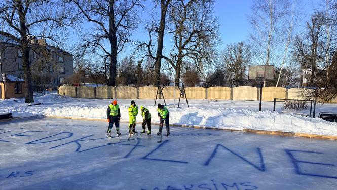 četri hokejistu komanda uz ledus ar nūjām, uz ledus liels uzraksts ZIEDKALNE