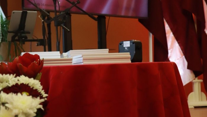 Priekšplānā sarkani un balti ziedi. Uz galda pasniedzamās pateicības. Fonā ekrāns ar Latvijas karogu