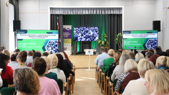 Skats no skatītāju zāles uz skatuvi ar ekrāniem un akmeņu attēliem