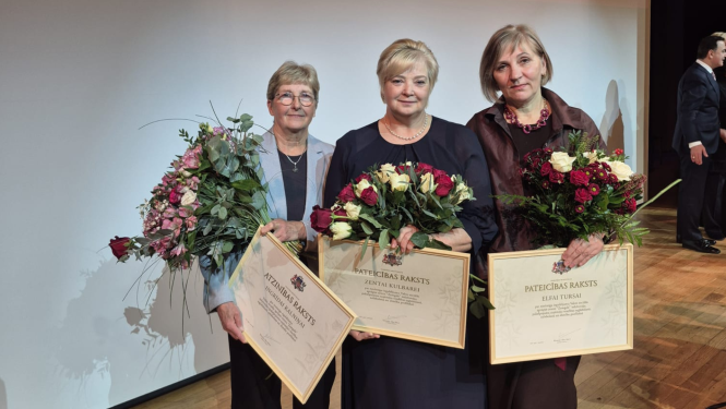 Fotogrāfijā trīs iestādes darbinieces ar ziediem un apbalvojumiem rokās