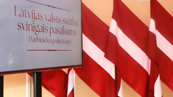 Attēlā ekrāns ar pasākuma baneri un fonā Latvijas karogu rinda