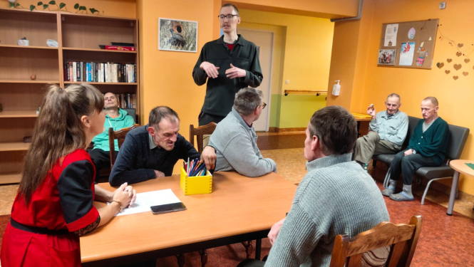 Attēlā darbiniece sarunājas ar iedzīvotājiem sēžot pie galda