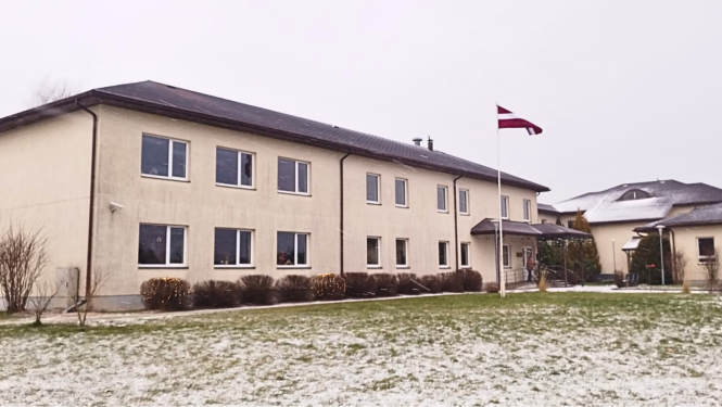 Grupu mājas divstāvu ēka bēšā krāsā, pie ieejas mastā plīvo valsts karogs