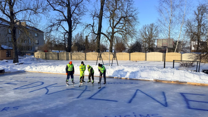 Četri hokejisti spilgti dzeltenajos tērpos stāv uz ledus ar nūjām. Uz ledus liels uzraksts ZIEDKALNE ar zilu krāsu
