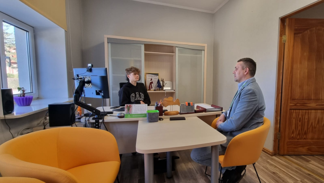 Kabinetā pie galda sēž iestādes direktors un jaunietis. Abi sarunājas.