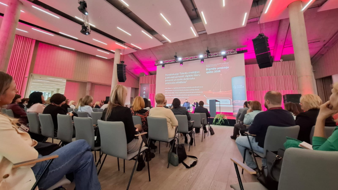 Fotogrāfija uzņemta Mežaparka Kokaru zālē. Attēlā konference, skats no skatītāju zāles, redzama pilna zāle ar konferences dalībniekiem, liels ekrāns un runātāji uz skatuves.