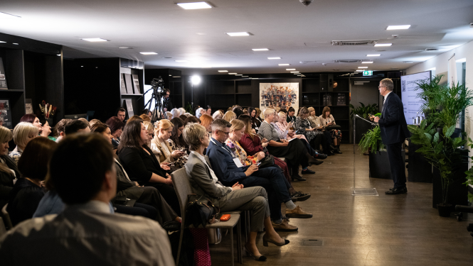 Konferences zāle pilna ar skatītājiem, kuri klausās vieslektora runā