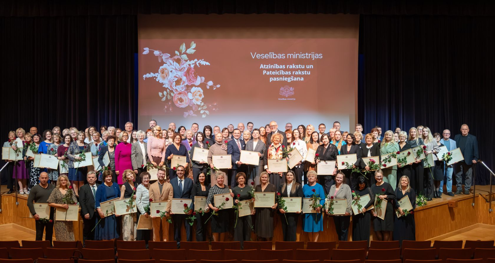 Kopbilde ar aptuveni simts cilvēkiem, kuri tur rokās ziedus un saņemtos ministrijas apbalvojumus
