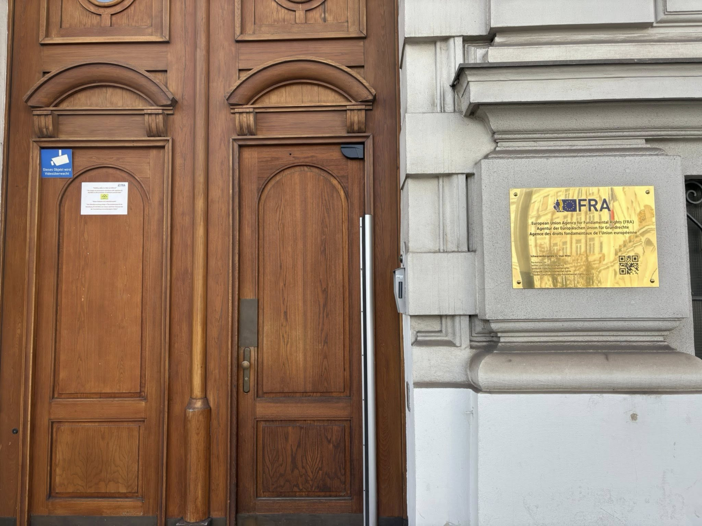 Lielas brūnas koka durvis un blakus iestādes informatīvā plāksne