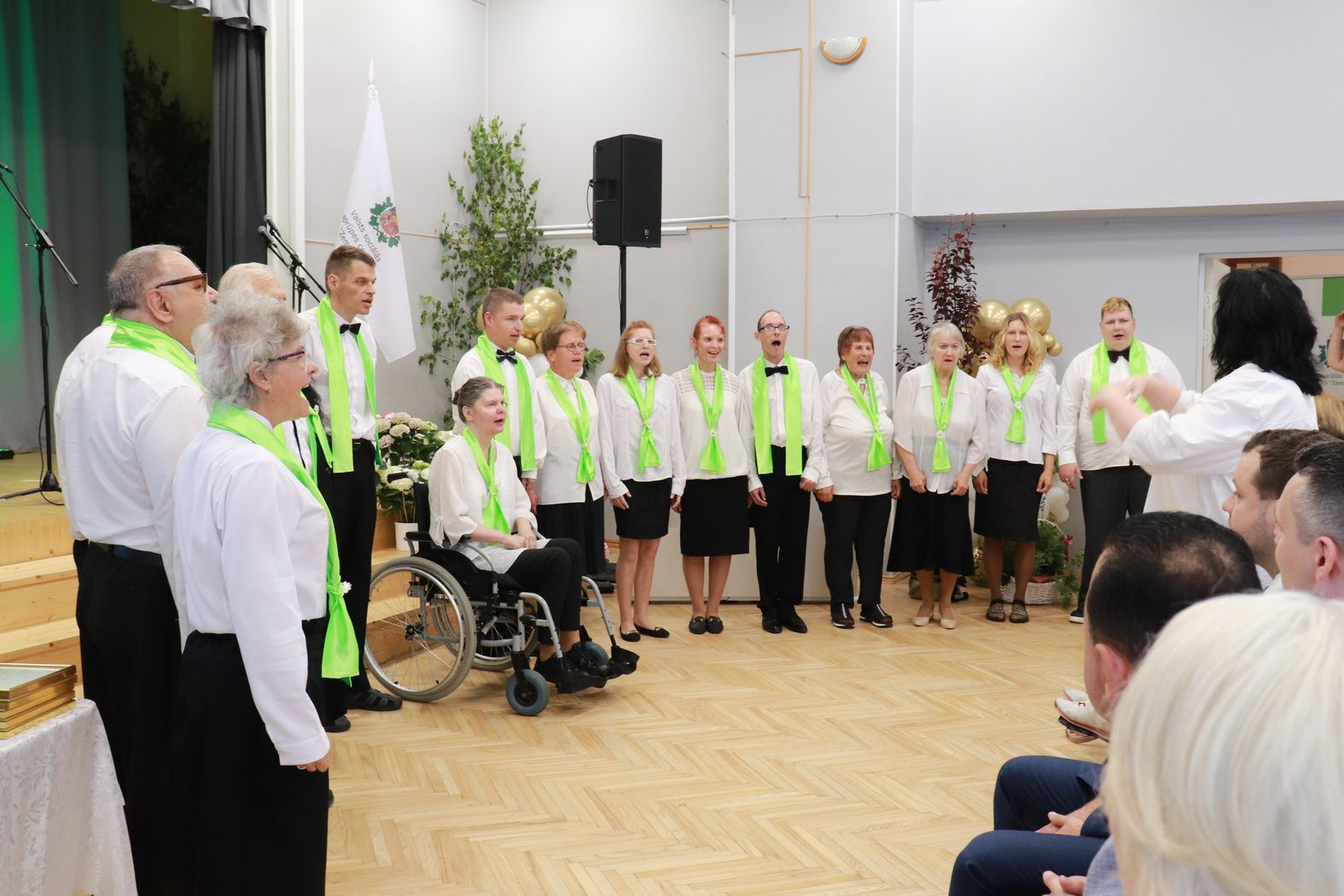 Uz skatuves iedzīvotāju apvienotais koris, baltajos kreklos, melnajās biksēs un visiem zaļas zīda šalles. Izpilda iestādes himnu.