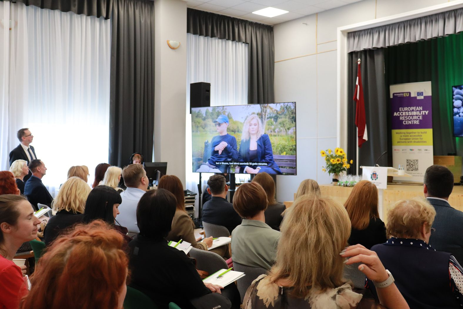 Domnīcas skatītāji sēž zālē un skatās uz ekrānā rādīto video