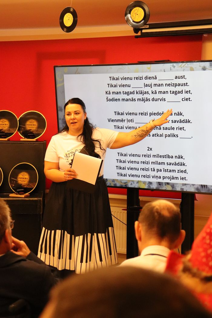 Pasākuma vadītāja pie ekrāna, kurā ir dziesmu minēšanas uzdevums