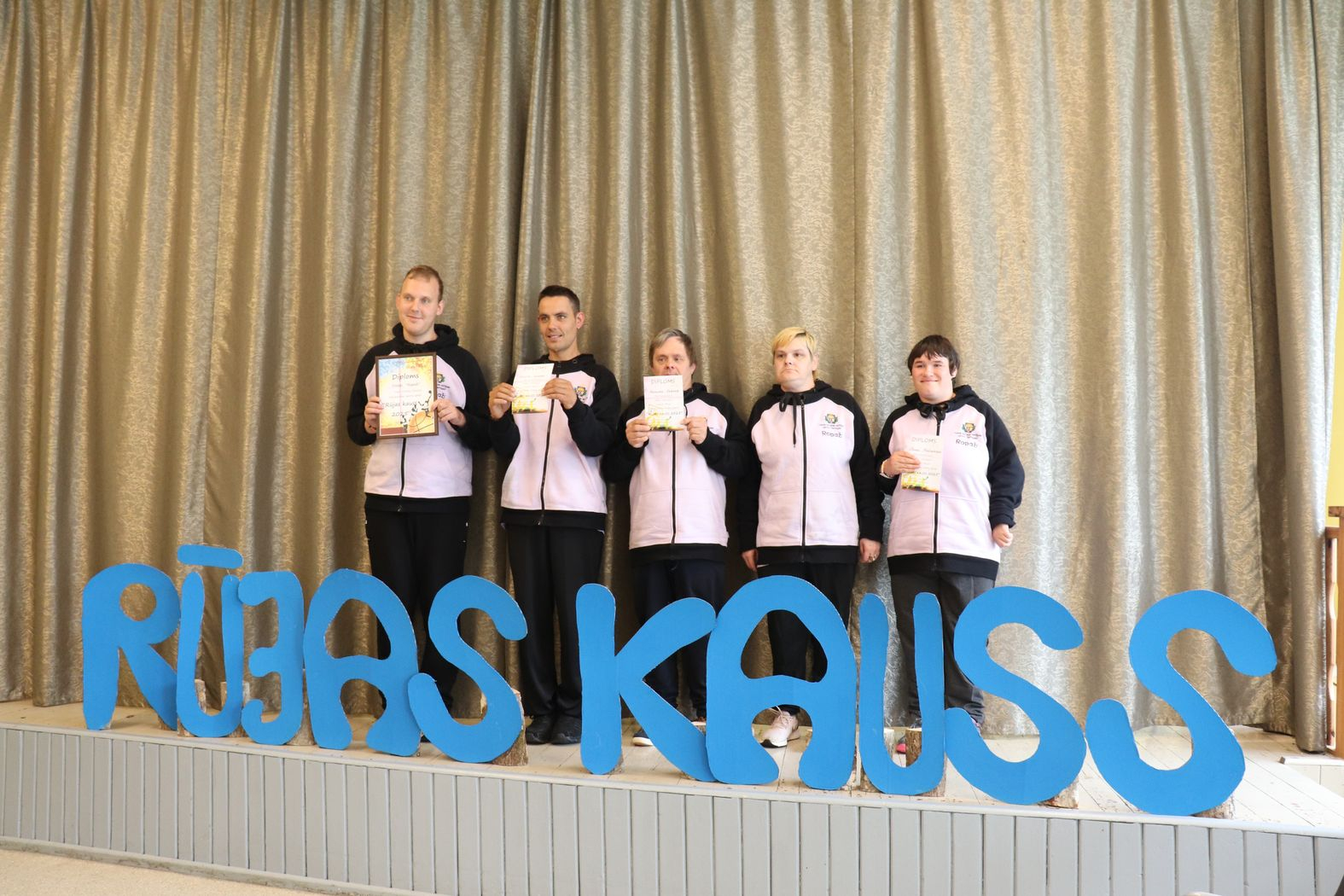 Komandas kopbilde ar diplomiem pie lielā uzraksta RŪJAS KAUSS
