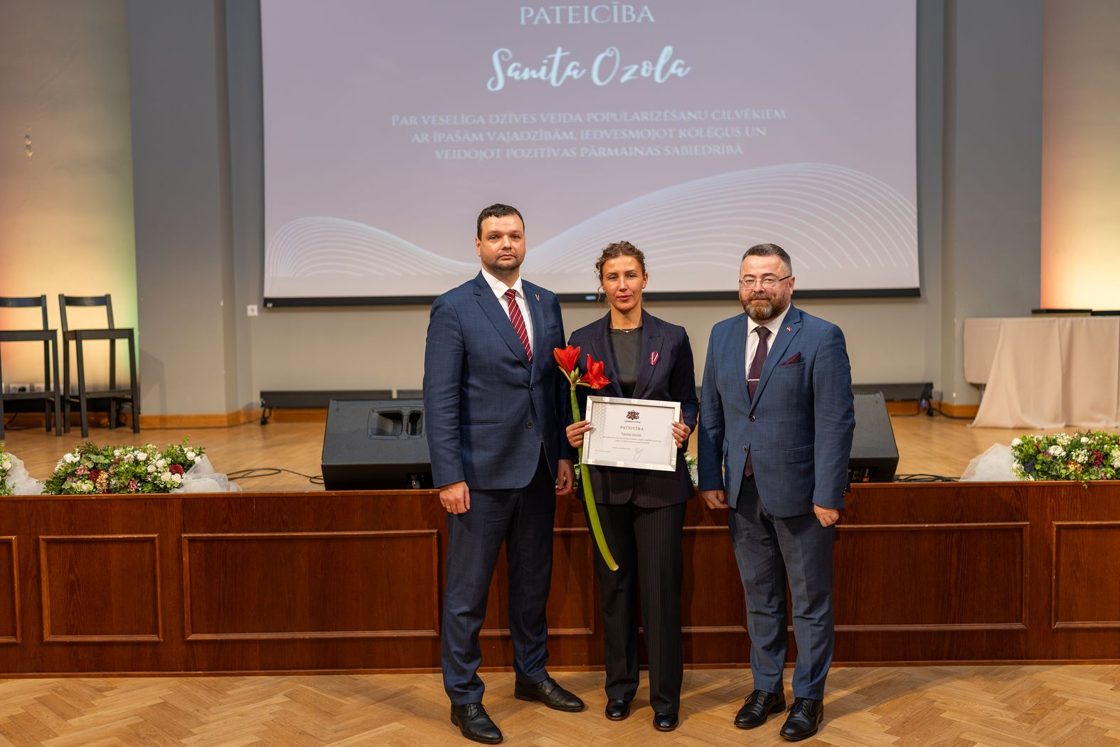 Apbalvojuma saņēmējs ar ziediem, pateicību kopbildē ar labklājības ministru un valsts sekretāru