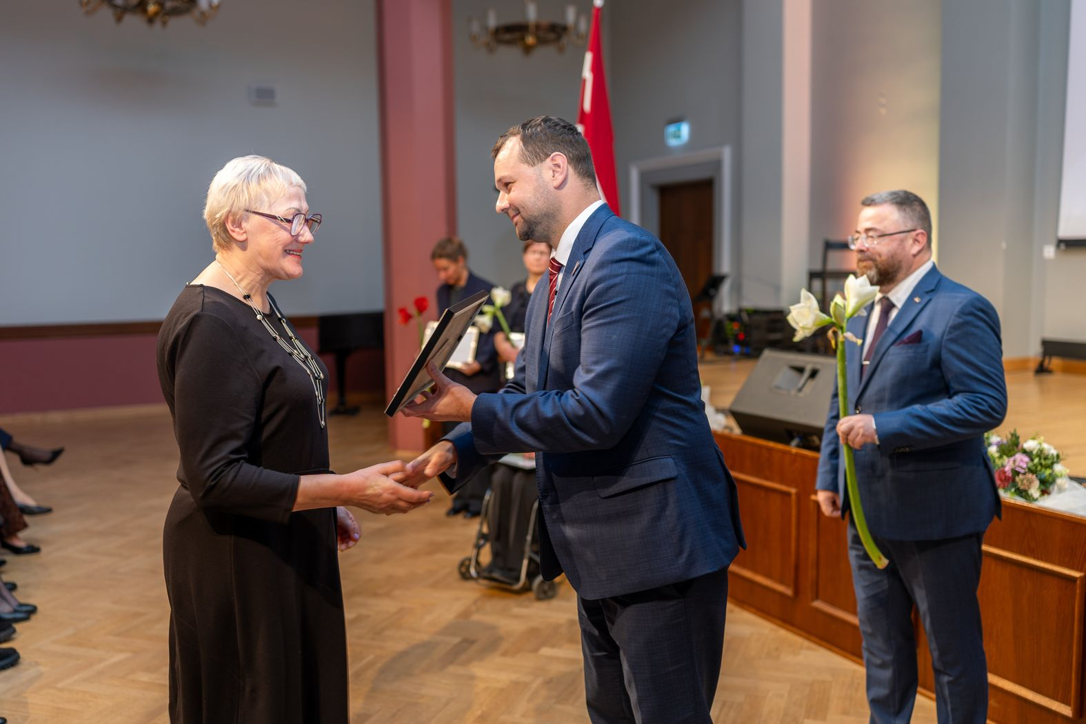 Apbalvojuma saņēmējs ar ziediem, pateicību kopbildē ar labklājības ministru un valsts sekretāru