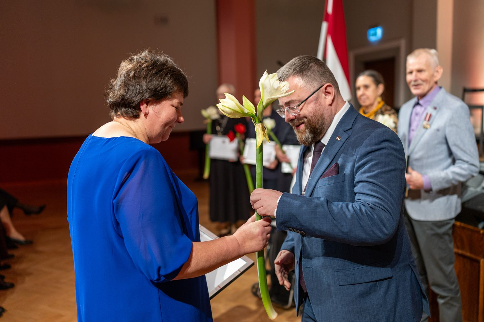 Apbalvojuma saņēmējs ar ziediem, pateicību kopbildē ar valsts sekretāru