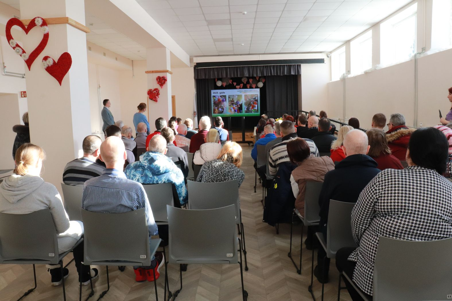 Zālē sēž iedzīvotāji, priekšā uz ekrāna tiek demonstrēta prezentācija