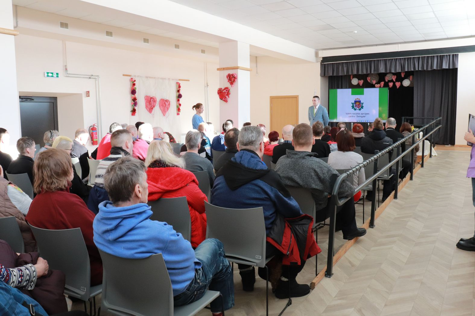 Zālē sēž iedzīvotāji, priekšā direktors vada kopsapulci