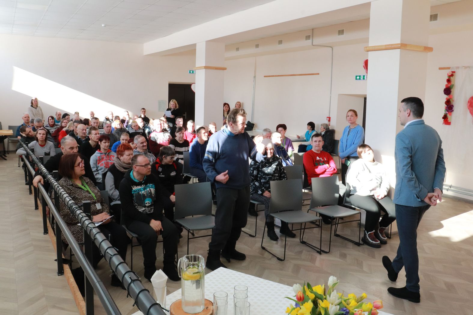 Zālē sēž iedzīvotāji, iedzīvotājs pirmajā rindā ir piecēlies kājās un uzdod direktoram jautājumu.