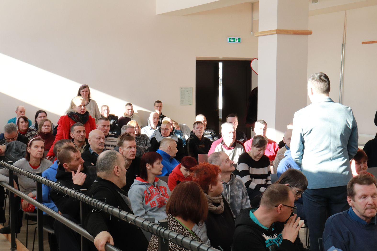 Zālē sēž iedzīvotāji, priekšā iestādes direktors vada kopsapulci, zālē kājās stāv iedzīvotāja sarkanā džemperī un uzdod jautājumu