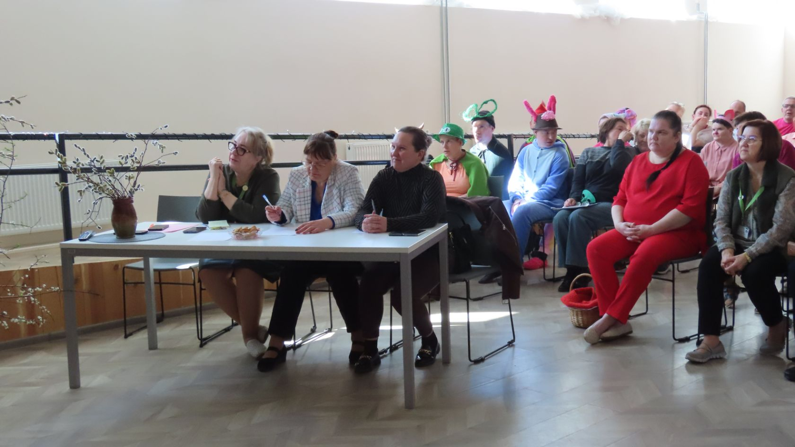 Pasākuma žūrija pie galda un pilna zāle ar skatītājiem