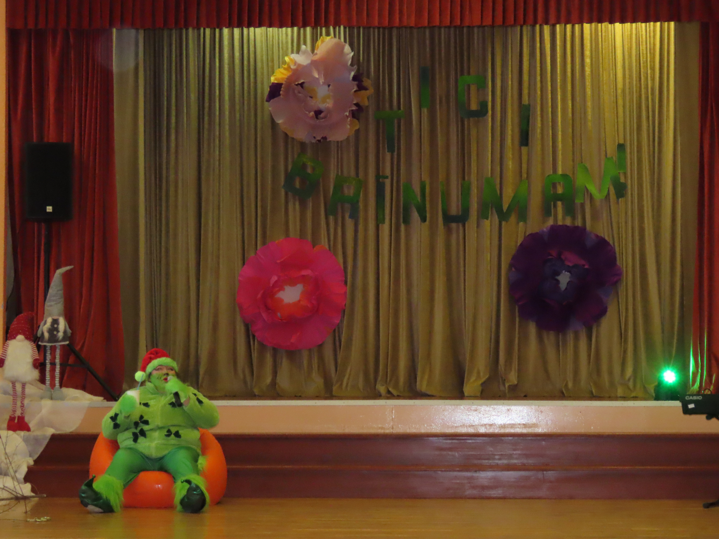 Skatuve ar uzrakstu TICI BRĪNUMAM un mīkstā oranžā pufa sēž darbiniece pārģērbusies Grinča tērpā.