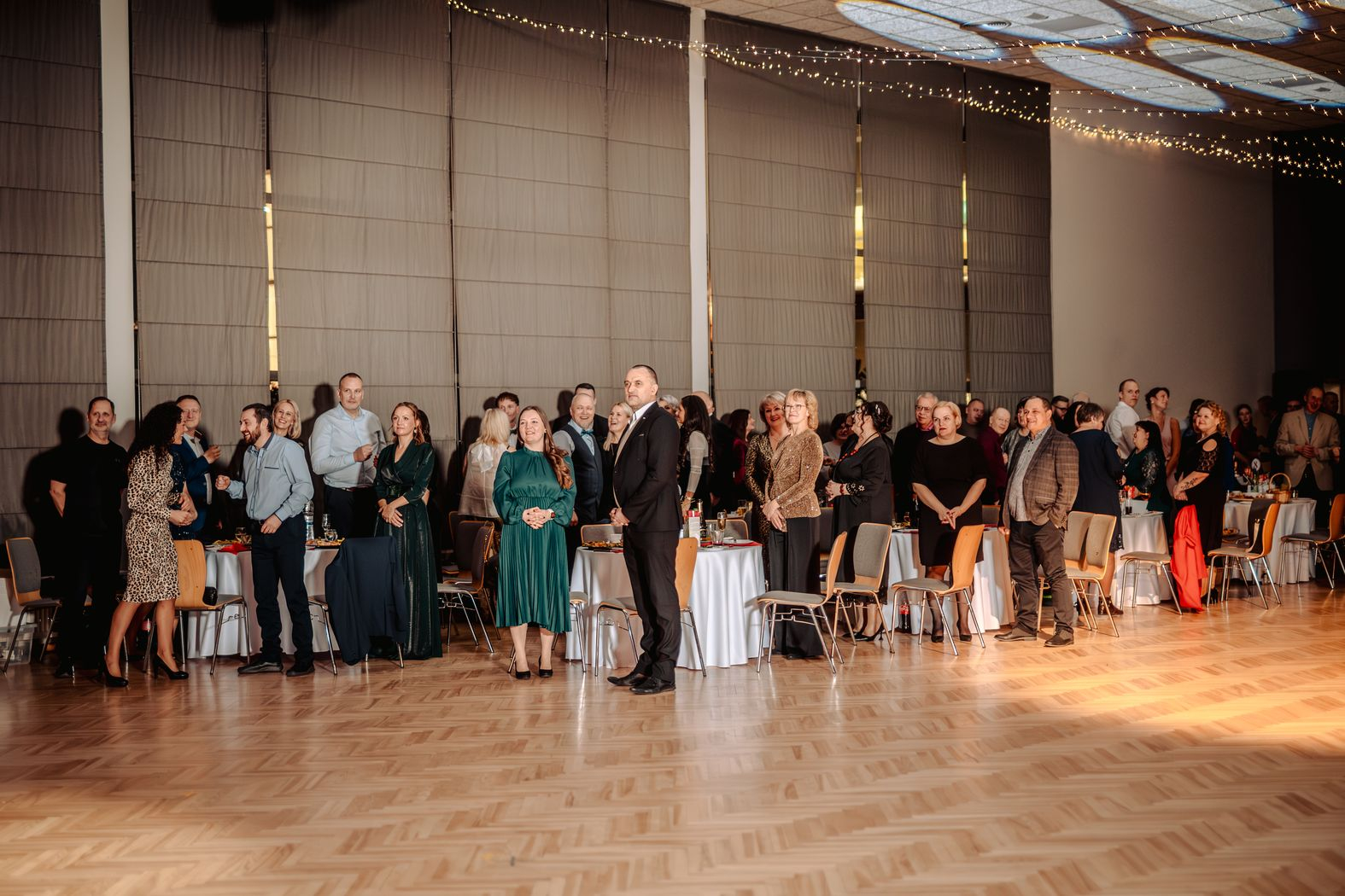 Pasākuma darbinieki pie galdiņiem stāv kājās