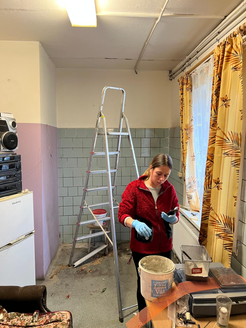 Telpa remonta veikšanas procesā. Netīras sienas baltā un lillā krāsā, blāvi zilas flīzes.