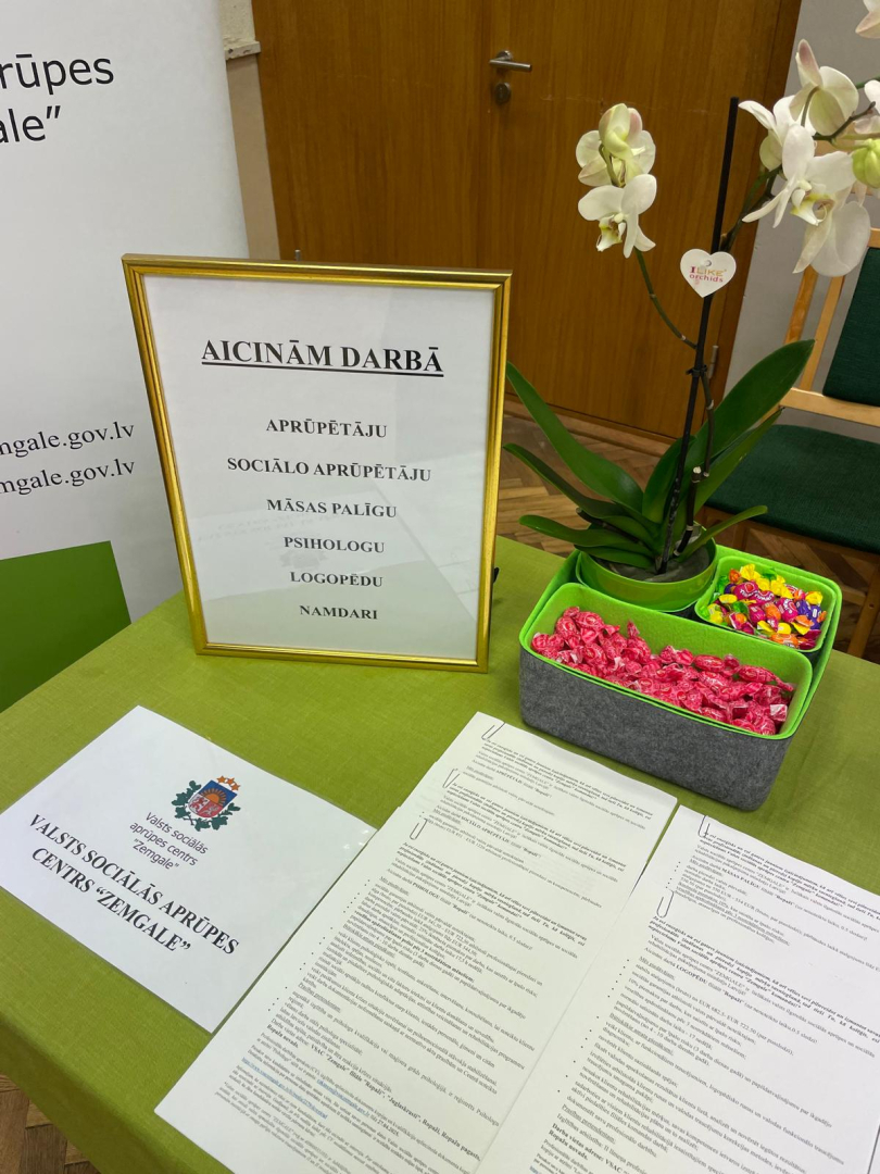 Uz galda novietoti dokumenti, konfektes, orhidejas zieds un VSAC "Zemgale" aktuālās vakances