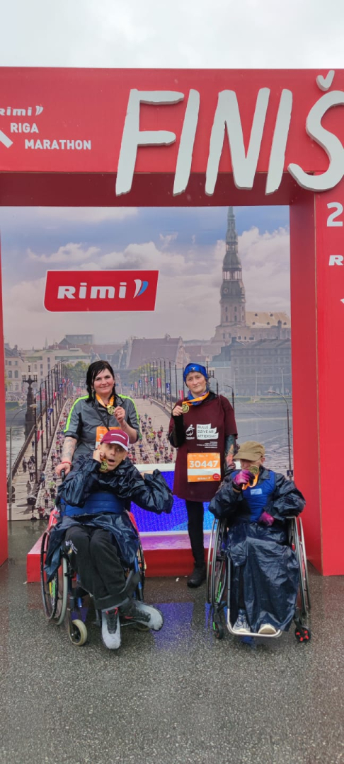 Rimi Rīgas maratona dalībnieki pēc distances veikšanas. Smaidīgi, ar medaļām. Divi iedzīvotāji ratiņkrēslos un divas pavadošās darbinieces