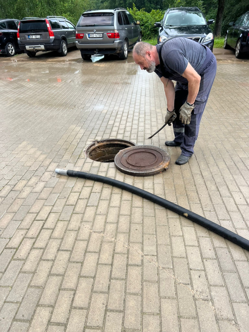 Attēlā āra kanalizācijas lūka, kurā darbinieks lej ūdeni