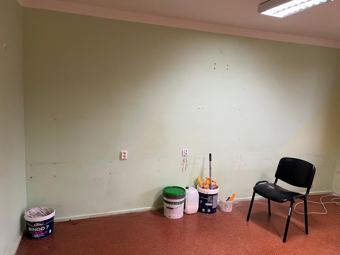 Telpa pirms remonta veikšanas. Nolietojušās sienas blāvi gaišā krāsā. Pie sienas remontdarbu materiāli, krāsas un citi