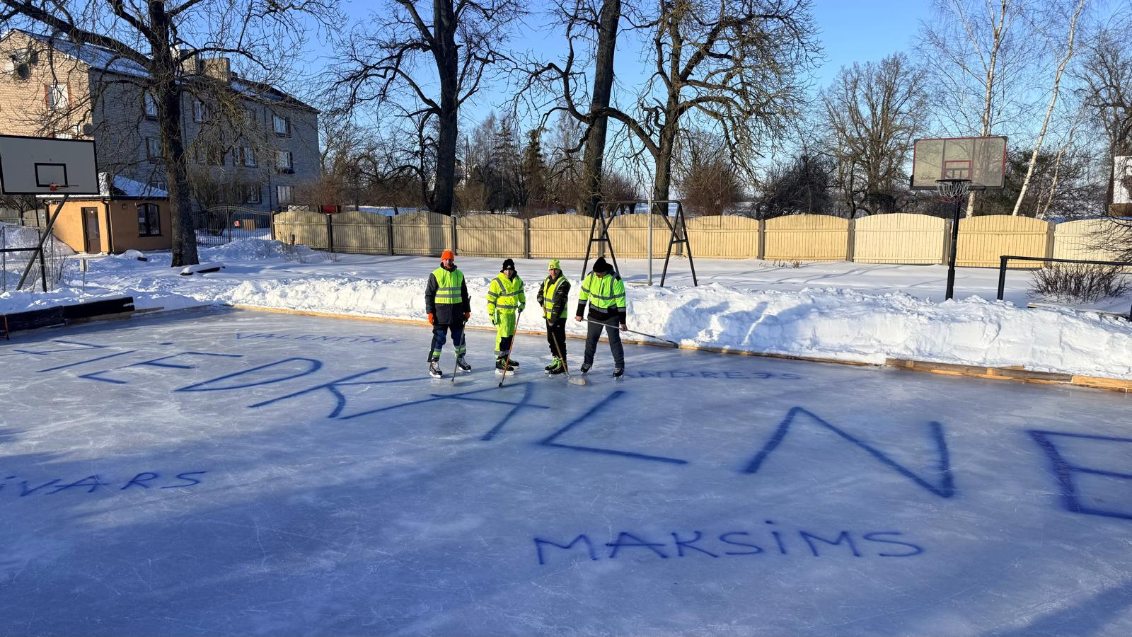 četri hokejistu komanda uz ledus ar nūjām, uz ledus liels uzraksts ZIEDKALNE