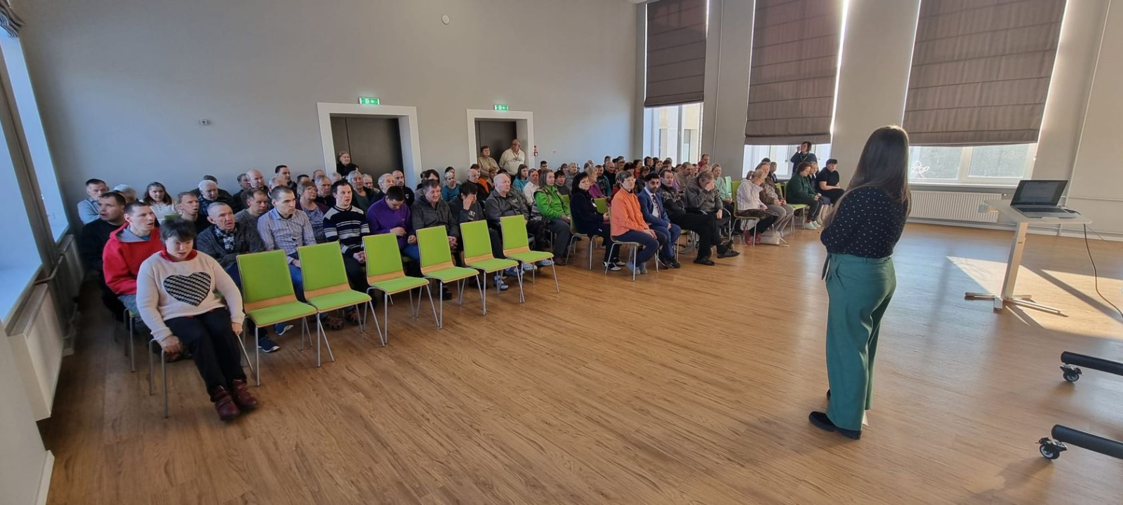 Zālē sēž iedzīvotāji, priekšā darbiniece zaļās biksēs un melnā blūzē vada kopsapulci