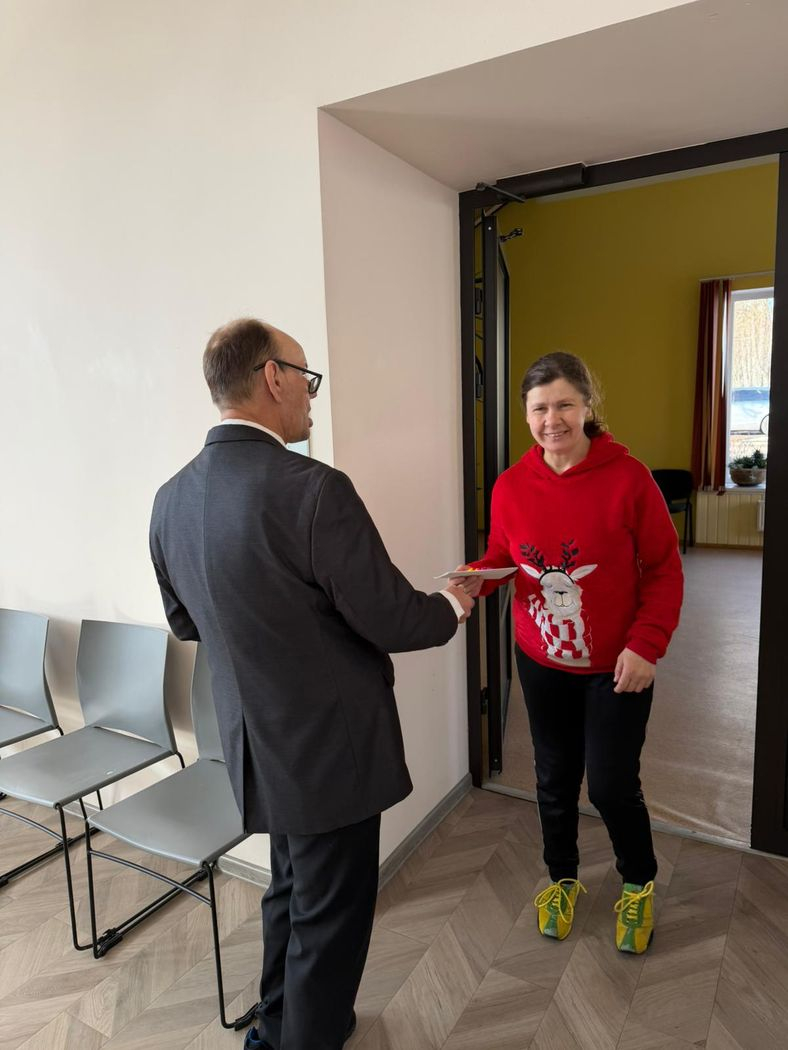 Kungs pasniedz apsveikuma kartīti dāmai, kura ienāk pa durvīm zālē