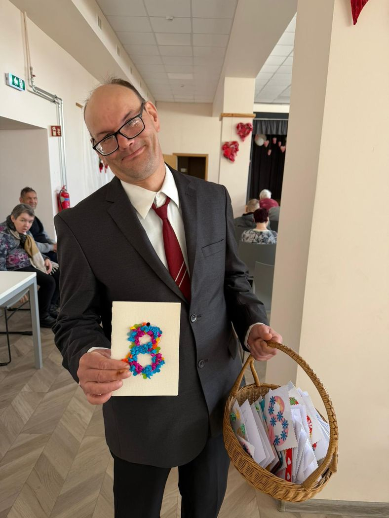 Smaidīgs kungs žaketē un ar brillēm tur rokā grozu ar apsveikumu kartītēm un otrā rokā kartīti ar ciparu 8