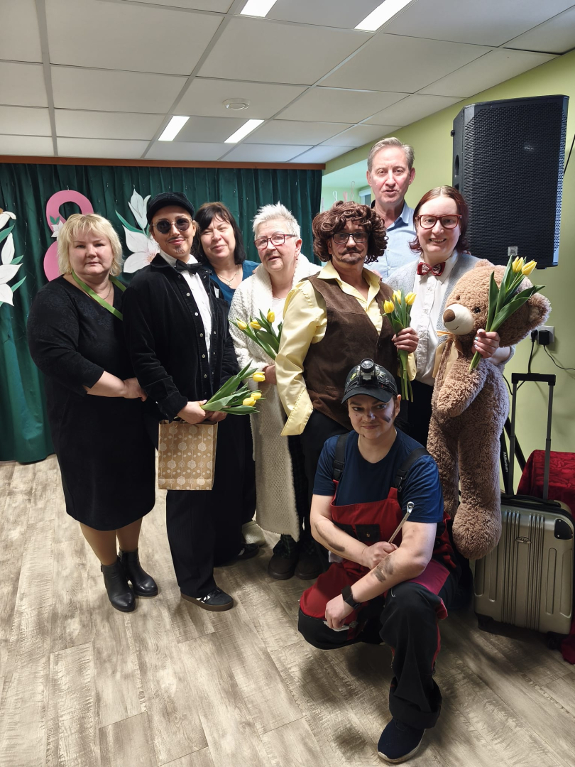 Attēlā astoņi cilvēki, filiāles darbinieki, tērpušies kostīmos un ar ziediem rokas