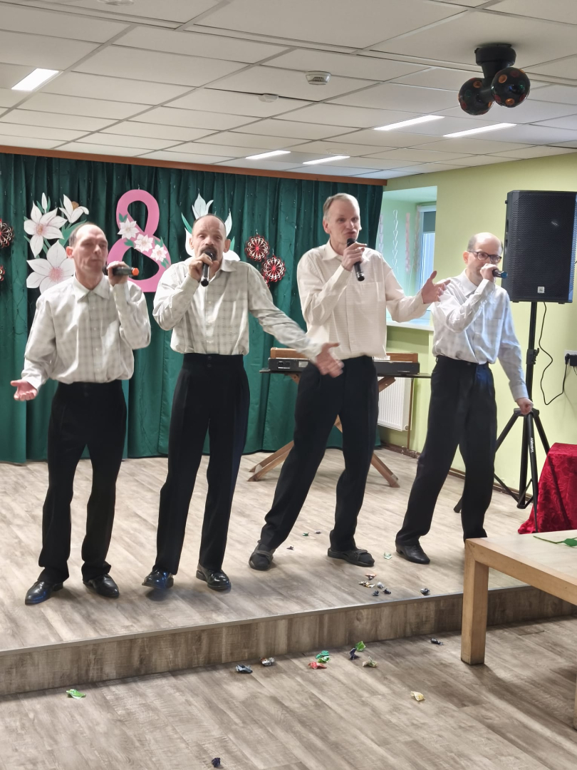 Attēlā četri kungi baltos kreklos un melnās biksēs. Stāv uz skatuves dzied turot rokās mikrofonus