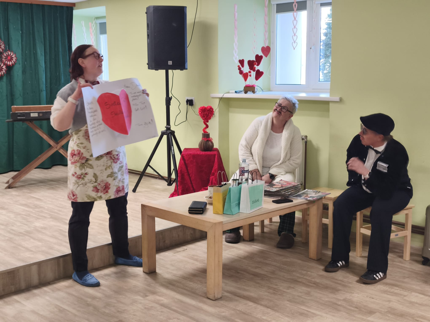 Trīs darbinieces rāda teātri, divas sēž krēslos, bet viena stāv turot lielu plakātu ar sarkanu sirdi uz tā